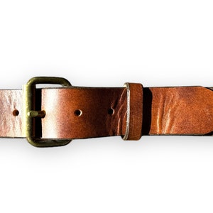 May include: Close-up of a brown leather belt with a brass buckle. The belt has several holes for adjustment and a pointed end. The leather shows natural variations in color and texture, with a warm, rustic appearance.