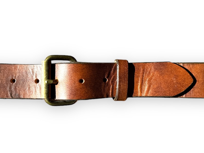 May include: Close-up of a brown leather belt with a brass buckle. The belt has several holes for adjustment and a pointed end. The leather shows natural variations in color and texture, with a warm, rustic appearance.