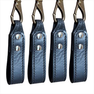 May include: Four black leather keychains with brass-colored clasps and silver rivets. Each keychain has a loop at the bottom and is stitched along the edges. The clasps are open.