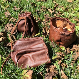May include: Three brown leather pouches with drawstrings. The pouches are sitting on a bed of green grass and brown leaves.