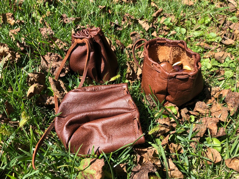 May include: Three brown leather pouches with drawstrings. The pouches are sitting on a bed of green grass and brown leaves.