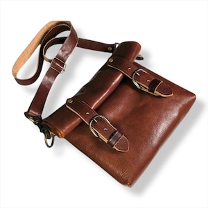 Könnte beinhalten: Eine braune Leder-Umhängetasche mit Schulterriemen und zwei Schnallenverschlüssen. Die Tasche hat eine rechteckige Form und eine Klappe, die sich über die Vorderseite legt. Der Riemen ist verstellbar.