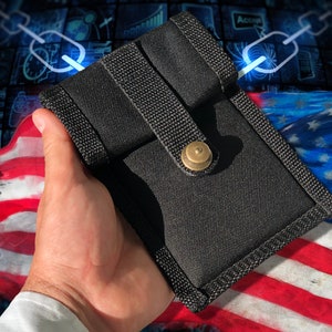 May include: A black fabric pouch with a flap closure and a brass button. The pouch is held in a hand against a background of an American flag.