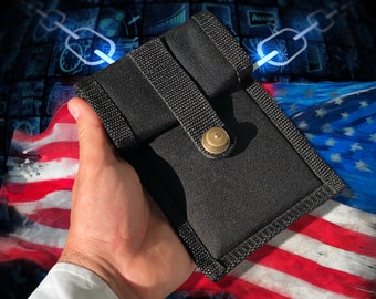 May include: A black fabric pouch with a flap closure and a brass button. The pouch is held in a hand against a background of an American flag.