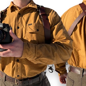 May include: A brown leather camera strap with a cross-body design. The strap is worn by a person wearing a yellow button-down shirt and brown pants. The strap has a metal buckle and a metal ring.