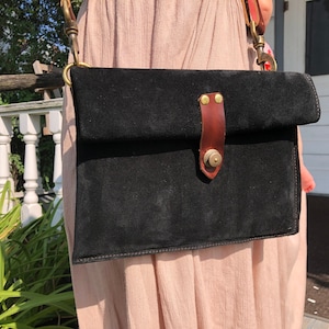 Könnte beinhalten: Eine schwarze Wildleder-Umhängetasche mit einem braunen Lederklappenverschluss und Messingbeschlägen. Die Tasche hat einen einzigen Riemen und einen Schnallenverschluss.