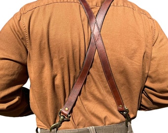 May include: Brown leather suspenders with brass hardware, worn over a brown button-down shirt and khaki pants.