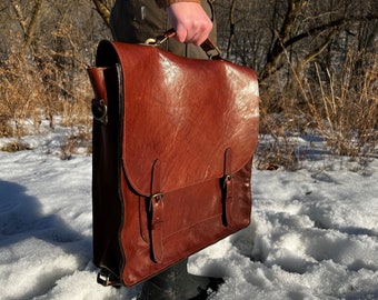 May include: A brown leather briefcase with two buckles and a strap. The briefcase is held by a hand in a snowy outdoor setting.