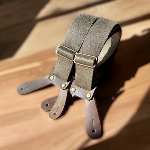Puede incluir: Un par de tirantes verde oliva con correas de cuero marrón y hebillas de latón. Los tirantes están hechos de tejido tejido y tienen un diseño clásico.