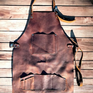 May include: A brown leather apron with multiple pockets and a large pocket on the front. The apron has adjustable straps and is made of thick, durable leather.