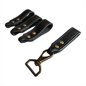 May include: Black leather key fobs with brass-colored rivets and stitching. One fob has a metal clasp and a square ring. Three additional key fobs are also shown.