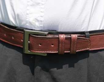 May include: A brown leather belt with a silver buckle. The belt is worn around the waist of a person wearing a white shirt and black pants.