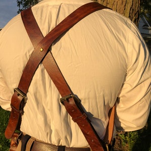 May include: Brown leather camera straps with brass buckles, crossing in an X pattern on the back of a person wearing a white shirt.
