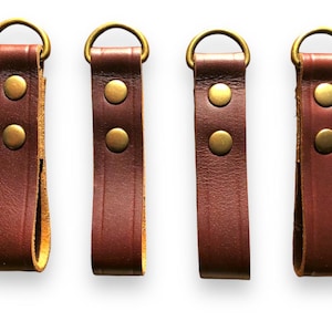 Pode incluir: Quatro tiras de couro marrom com argolas de latão e dois rebites de latão cada. As tiras medem aproximadamente 5 cm de comprimento.