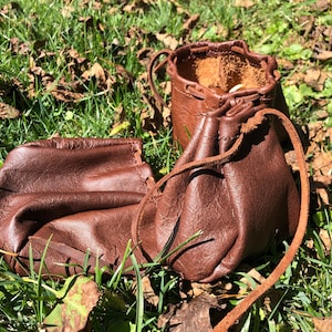 May include: Two brown leather pouches with drawstrings, lying on a bed of green grass and brown leaves.