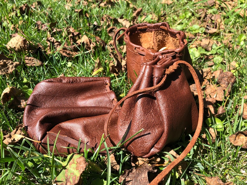 May include: Two brown leather pouches with drawstrings, lying on a bed of green grass and brown leaves.