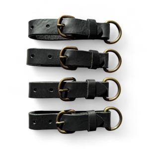 Op de afbeelding: Vier zwarte leren sleutelhangers met messingkleurige gespen en cirkelvormige ringen. De sleutelhangers zijn horizontaal geplaatst tegen een witte achtergrond. Elke sleutelhanger heeft een gesp en een ring om sleutels aan te bevestigen.