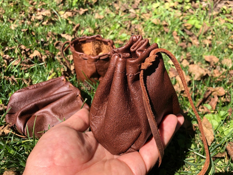 May include: Two brown leather pouches with drawstrings. One pouch is open and the other is closed. The pouches are sitting on a bed of green grass.