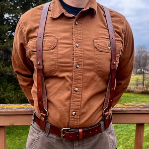 Può includere: Una persona indossa bretelle in pelle marrone con una cintura in pelle marrone e pantaloni marroni. Le bretelle hanno fibbie in ottone e la cintura ha una fibbia in ottone.