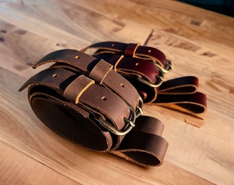 May include: Three leather belts with brass buckles. The belts are brown and have a double layer design. The belts are arranged in a pile on a wooden surface.