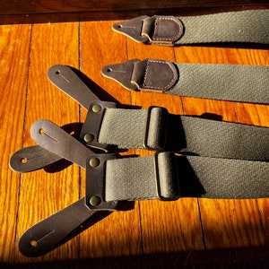 May include: A pair of green canvas suspenders with brown leather straps and brass buckles. The suspenders are laid out flat on a wooden surface.