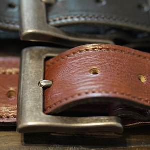 May include: Close-up of a brown leather belt with a silver buckle. The belt has stitching along the edges and two rows of holes.