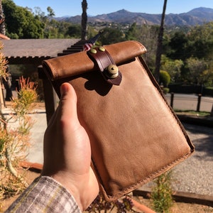 May include: A brown leather phone case with a brown leather strap and a brass buckle. The case has a flap that folds over the top of the phone.