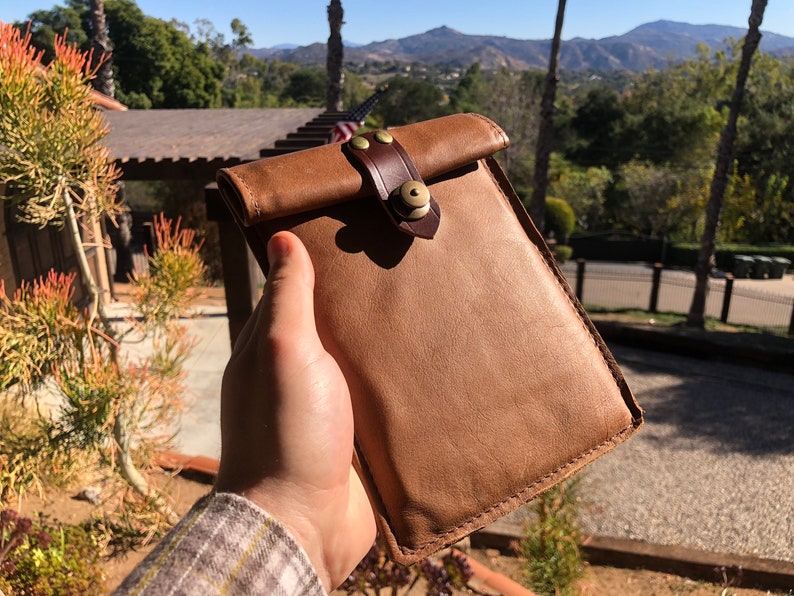 May include: A brown leather phone case with a brown leather strap and a brass buckle. The case has a flap that folds over the top of the phone.