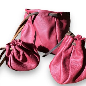 May include: Three red leather pouches of varying sizes. The pouches have a drawstring closure and brown leather straps. The leather has a textured appearance. The pouches are likely for carrying small items.