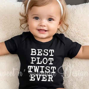 May include: A smiling baby wearing a black onesie with the text "BEST PLOT TWIST EVER" in white. The baby has a white bow headband and is sitting on a fluffy white pillow.
