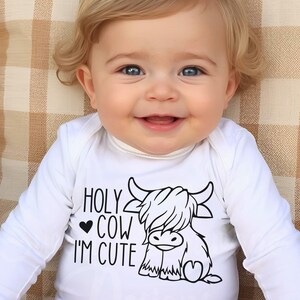 May include: A white long-sleeved baby bodysuit featuring a black graphic print of a Highland cow and the text "HOLY COW I'M CUTE" with a small heart symbol. The garment has a simple crew neckline and long sleeves, made of a plain white fabric.