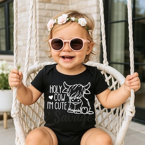 May include: A black short-sleeved baby bodysuit featuring a white graphic of a Highland cow and the text "HOLY COW I'M CUTE" with a small heart icon. The garment has a simple crew neckline and is made of a solid black fabric with white ink.