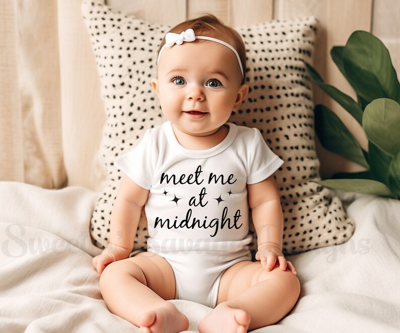 May include: A baby wearing a white onesie with the text "meet me at midnight" and a white headband with a bow. The baby is sitting on a white blanket with a brown and white patterned pillow behind them.