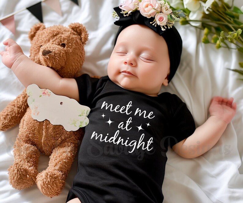 May include: A baby wearing a black onesie with the text "Meet me at midnight" in white. The baby is sleeping and has a flower headband on. A brown teddy bear is next to the baby.