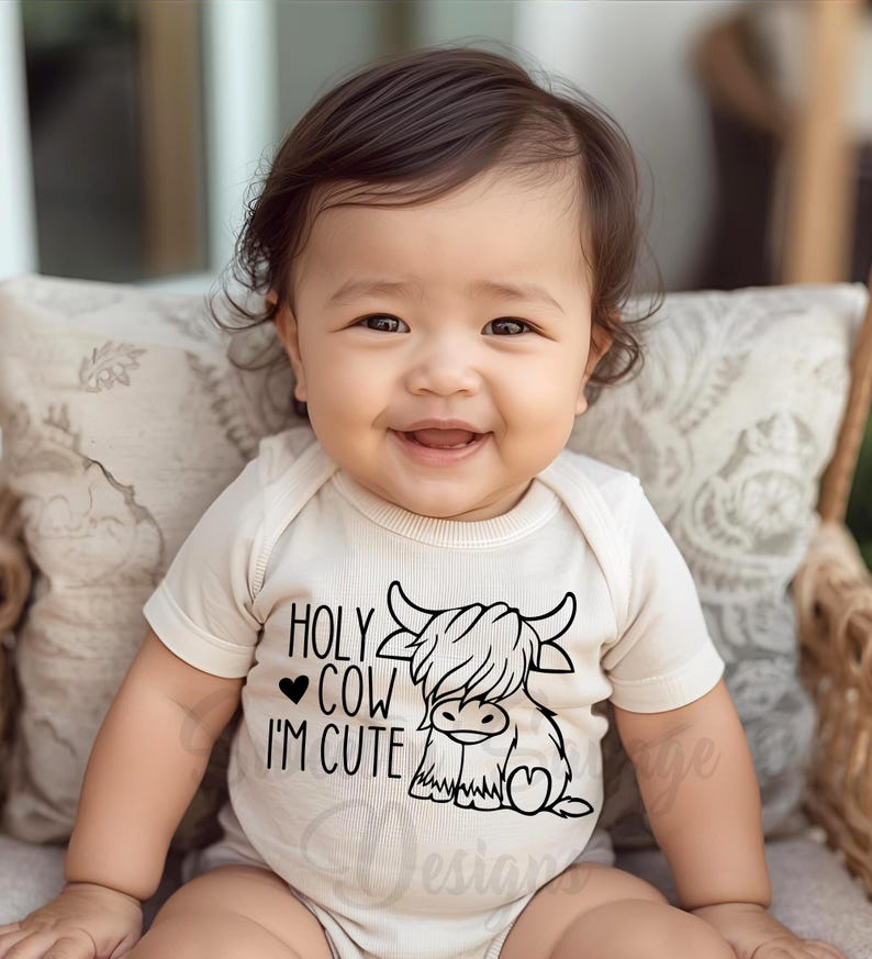 May include: A cream-colored short-sleeve baby bodysuit featuring a black graphic of a Highland cow and the text "HOLY COW I'M CUTE" with a small heart symbol. The garment has a ribbed texture and a simple round neckline for easy dressing.