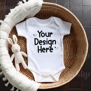 May include: A white baby onesie with the text "Your Design Here" in black, placed in a woven basket. A white stuffed bunny and a white knitted blanket are also in the basket. The basket is on a dark brown wooden surface.