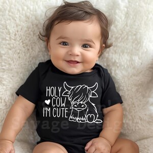 May include: A black short-sleeved baby bodysuit featuring a white graphic of a Highland cow and the text "HOLY COW I'M CUTE" with a small heart icon. The design is printed on the front of the garment, which has a simple round neckline.