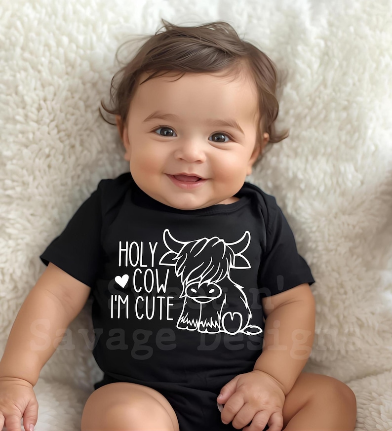 May include: A black short-sleeved baby bodysuit featuring a white graphic of a Highland cow and the text "HOLY COW I'M CUTE" with a small heart icon. The design is printed on the front of the garment, which has a simple round neckline.