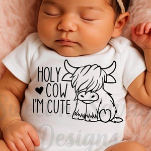 May include: A white short-sleeved baby bodysuit featuring a black graphic print of a Highland cow and the text "HOLY COW I'M CUTE" with a small heart icon. The design is centered on the chest of the garment, which is worn by a sleeping infant.