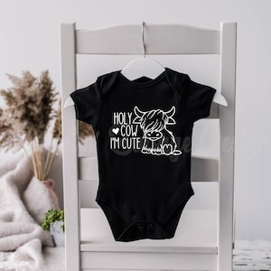 May include: A black short-sleeved baby bodysuit featuring a white graphic print of a Highland cow. The text "HOLY COW I'M CUTE" is printed in white lettering next to the cow illustration, which includes a small heart icon near the text.