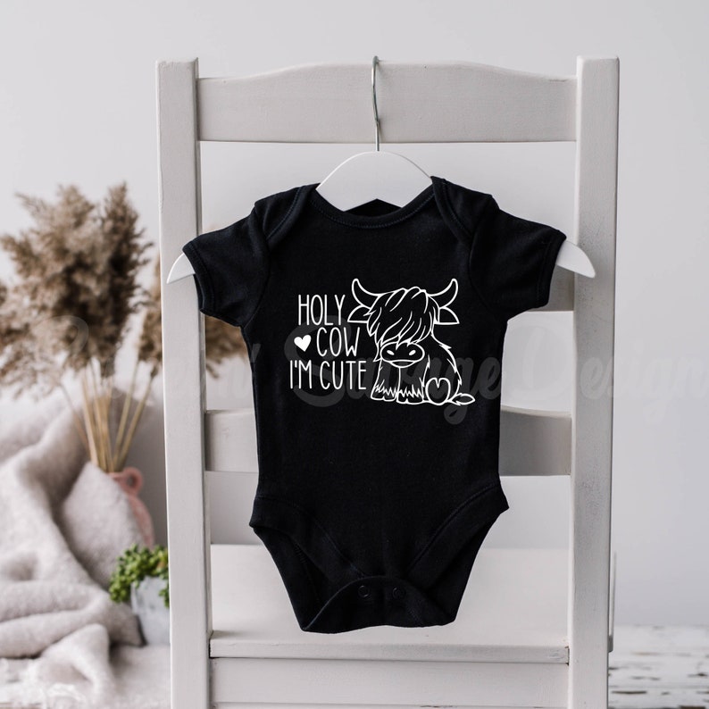 May include: A black short-sleeved baby bodysuit featuring a white graphic print of a Highland cow. The text "HOLY COW I'M CUTE" is printed in white lettering next to the cow illustration, which includes a small heart icon near the text.