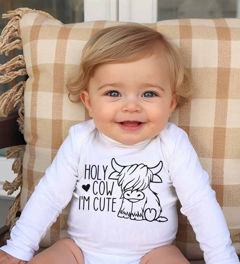 May include: A white long-sleeved baby bodysuit featuring a black graphic print of a Highland cow and the text "HOLY COW I'M CUTE" with a small heart symbol. The garment has a simple crew neckline and long sleeves, made of a plain white fabric.