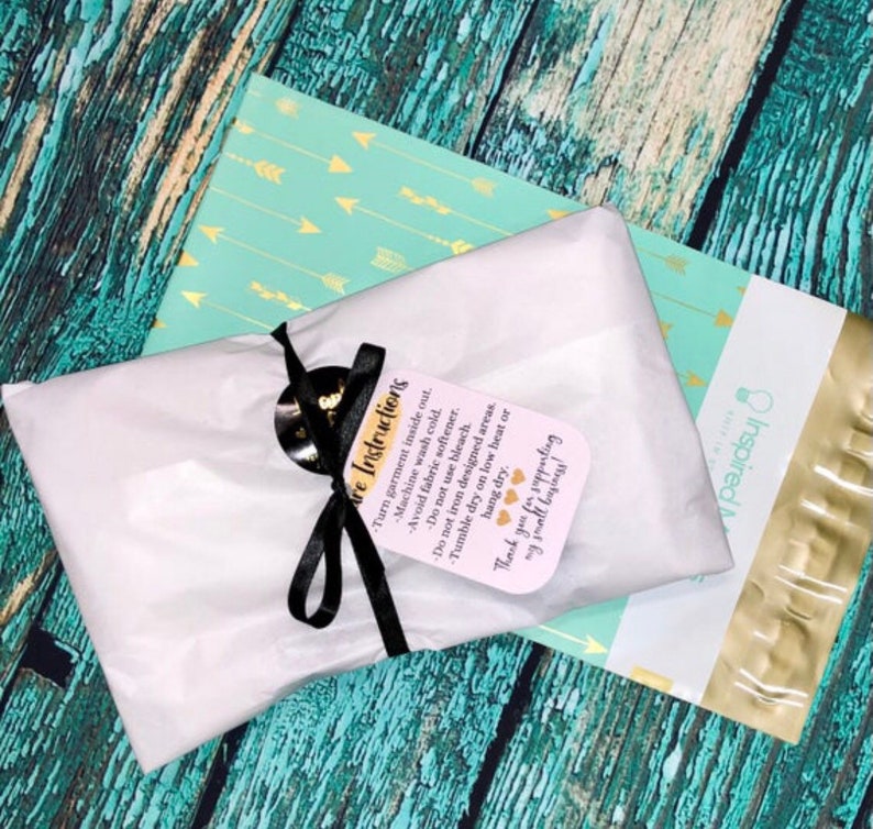 May include: A rectangular package wrapped in white tissue paper, tied with a black ribbon and a tag that reads: Care Instructions. Turn garment inside out. Machine wash cold. Avoid fabric softener. Do not use bleach. Do not iron designed areas. Tumble dry on low heat or hang dry. Thank you for supporting my small business!