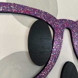 Puede incluir: Primer plano de una pieza de arte mural decorativa con un par de gafas moradas de gran tama&ntilde;o cubiertas de purpurina. Las gafas est&aacute;n colocadas sobre un fondo de formas blancas y grises, con formas ovaladas negras que representan los ojos.