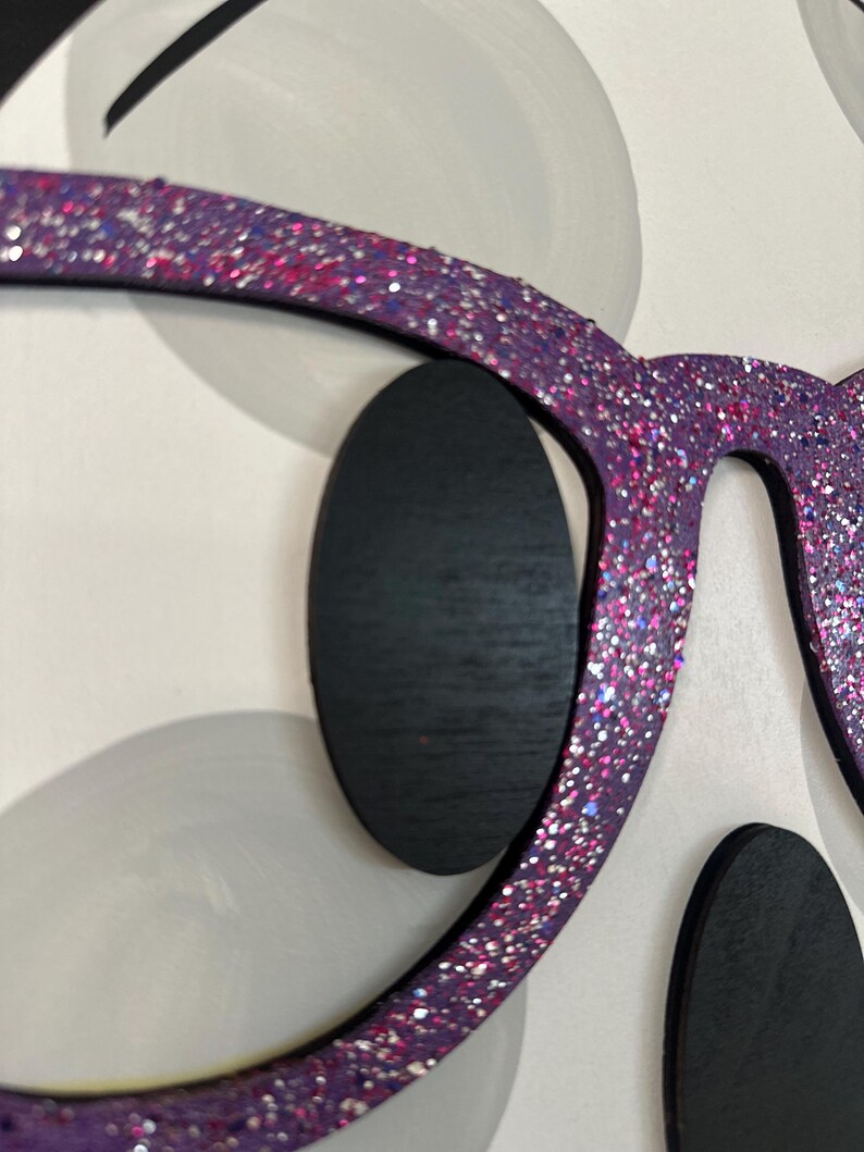 Puede incluir: Primer plano de una pieza de arte mural decorativa con un par de gafas moradas de gran tama&ntilde;o cubiertas de purpurina. Las gafas est&aacute;n colocadas sobre un fondo de formas blancas y grises, con formas ovaladas negras que representan los ojos.
