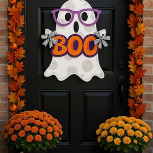 Puede incluir: Una puerta principal negra decorada para Halloween con un fantasma blanco con gafas moradas y la palabra "BOO" en naranja. La puerta est&aacute; flanqueada por follaje de oto&ntilde;o y calabazas, con crisantemos naranjas en macetas debajo.