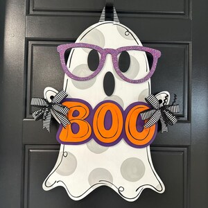Puede incluir: Decoraci&oacute;n de fantasma de Halloween de madera con gafas de purpurina moradas y la palabra "BOO" en letras naranjas. El fantasma tiene lunares grises y lazos a rayas blancas y negras a cada lado. Cuelga de una puerta negra.