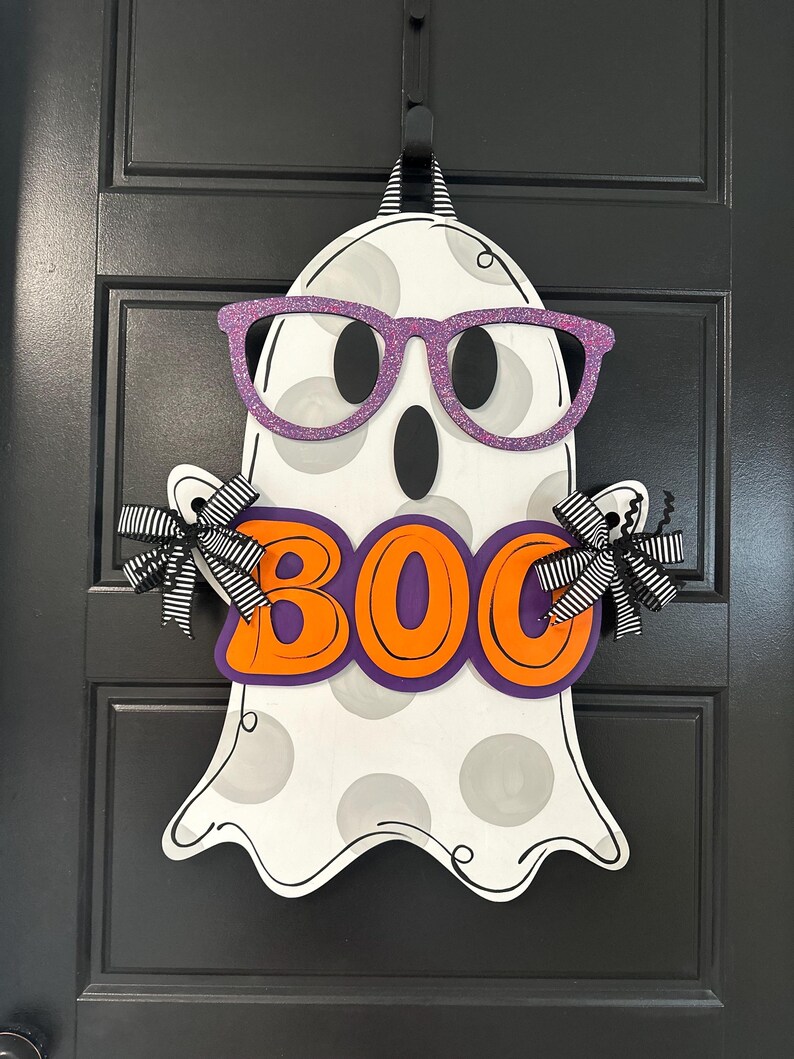 Puede incluir: Decoraci&oacute;n de fantasma de Halloween de madera con gafas de purpurina moradas y la palabra "BOO" en letras naranjas. El fantasma tiene lunares grises y lazos a rayas blancas y negras a cada lado. Cuelga de una puerta negra.