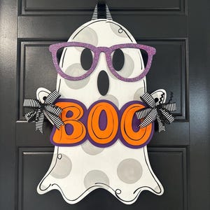 Puede incluir: Un colgador de puerta de Halloween con forma de fantasma de madera. El fantasma es blanco con lunares grises, gafas de purpurina moradas y la palabra "BOO" en letras naranjas con un contorno morado. Lazos a rayas blancas y negras est&aacute;n unidos a los brazos del fantasma.