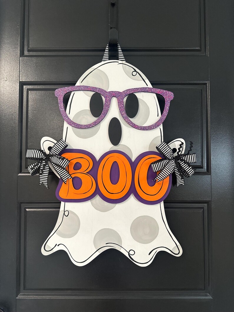 Puede incluir: Un colgador de puerta de Halloween con forma de fantasma de madera. El fantasma es blanco con lunares grises, gafas de purpurina moradas y la palabra "BOO" en letras naranjas con un contorno morado. Lazos a rayas blancas y negras est&aacute;n unidos a los brazos del fantasma.